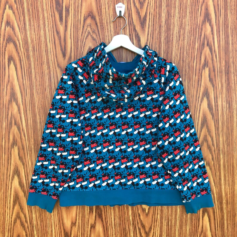 May include: A teal hoodie with a repeating pattern of Mickey Mouse faces in red, black, and white. The hoodie has a hood, a drawstring, and teal cuffs and hem. The garment is hanging on a white hanger against a wooden background.