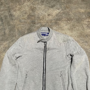 May include: A light gray zip-up jacket with a stand-up collar. The jacket has a full-length zipper, two side pockets, and the word "MAN" printed on the collar. The jacket is made of a soft, textured material.