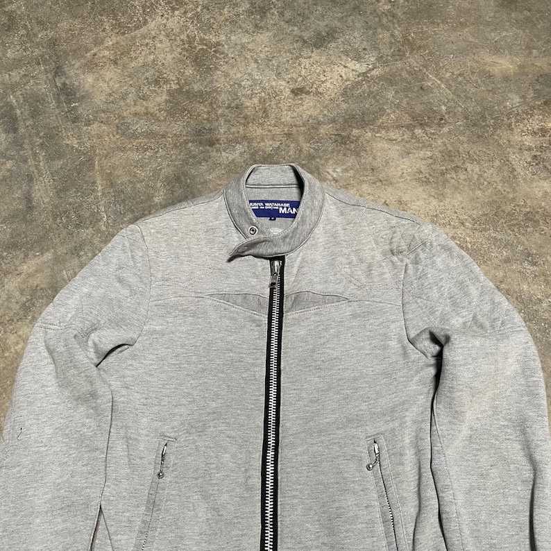 May include: A light gray zip-up jacket with a stand-up collar. The jacket has a full-length zipper, two side pockets, and the word "MAN" printed on the collar. The jacket is made of a soft, textured material.