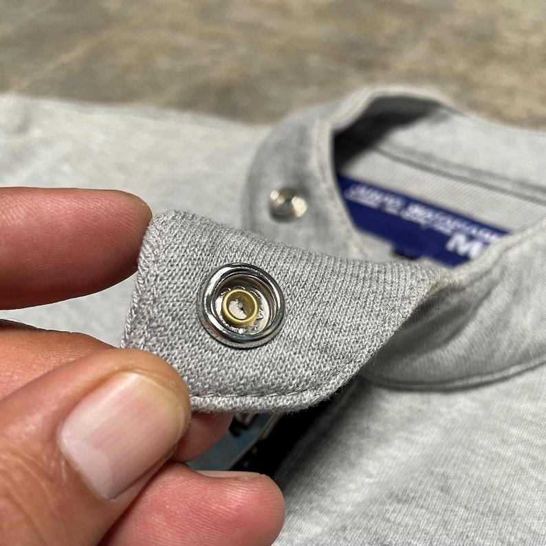 May include: Close-up of a gray fabric with a metal snap closure. The snap has a silver rim and a gold center. The fabric appears to be a soft knit material, possibly cotton or a blend. The image is well-lit, highlighting the details of the snap.