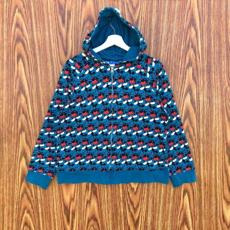 May include: Teal zip-up hoodie with a repeating pattern of Mickey Mouse faces in red, black, and white. The hoodie features a matching teal hood, cuffs, and waistband, and a full-length zipper.