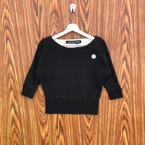 Könnte beinhalten: Schwarzes Sweatshirt mit Dreiviertelärmeln und weitem Ausschnitt. Auf der rechten Seite befindet sich ein weißes Blumenemblem. Der Markenname "MARY QUANT LONDON" ist im Innenausschnitt aufgedruckt.