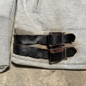 May include: Close-up of a gray sweatshirt with black leather straps and silver buckles. The sweatshirt has a zipper at the neckline and ribbed cuffs. The leather straps are buckled across the bottom of the sweatshirt.