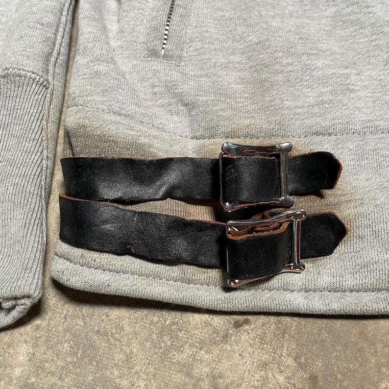 May include: Close-up of a gray sweatshirt with black leather straps and silver buckles. The sweatshirt has a zipper at the neckline and ribbed cuffs. The leather straps are buckled across the bottom of the sweatshirt.