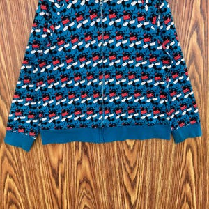 May include: A blue zip-up hoodie featuring a repeating pattern of Mickey Mouse. The design includes red, black, and white elements. The cuffs and hem are a solid teal color. The hoodie has a full-length zipper.