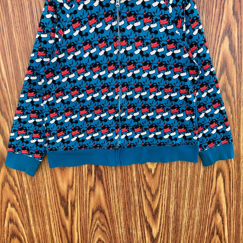 May include: A blue zip-up hoodie featuring a repeating pattern of Mickey Mouse. The design includes red, black, and white elements. The cuffs and hem are a solid teal color. The hoodie has a full-length zipper.