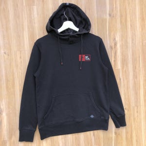 May include: Black hooded sweatshirt with a kangaroo pocket and the Rip Curl logo on the chest. The sweatshirt has a drawstring hood and button details at the neck. The garment is displayed on a wooden surface.