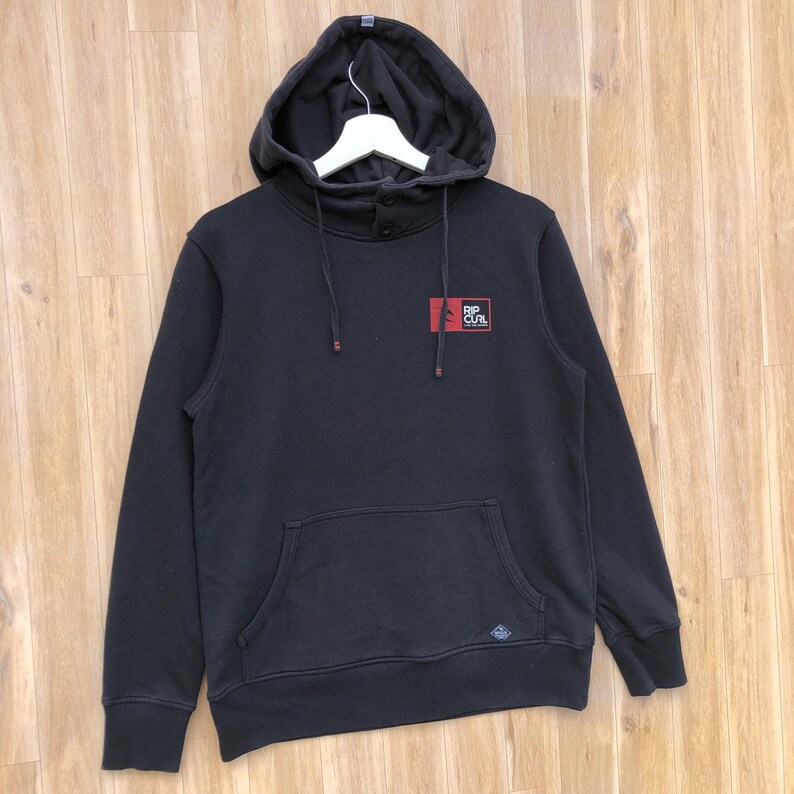 May include: Black hooded sweatshirt with a kangaroo pocket and the Rip Curl logo on the chest. The sweatshirt has a drawstring hood and button details at the neck. The garment is displayed on a wooden surface.