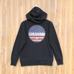 May include: Black hooded sweatshirt with a circular graphic on the back featuring a blue field of stars and red and white stripes, with the brand name "RIP CURL" in white. The sweatshirt has a drawstring hood.