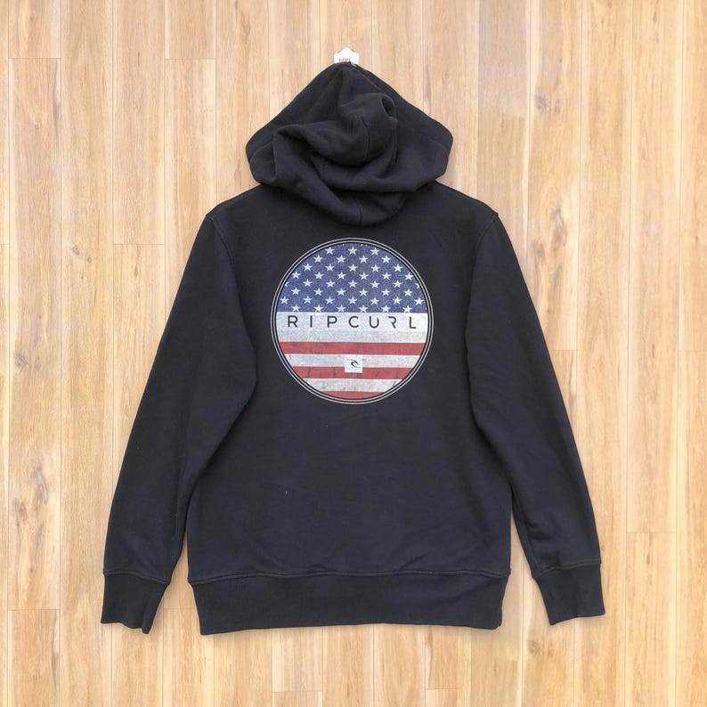 May include: Black hooded sweatshirt with a circular graphic on the back featuring a blue field of stars and red and white stripes, with the brand name "RIP CURL" in white. The sweatshirt has a drawstring hood.