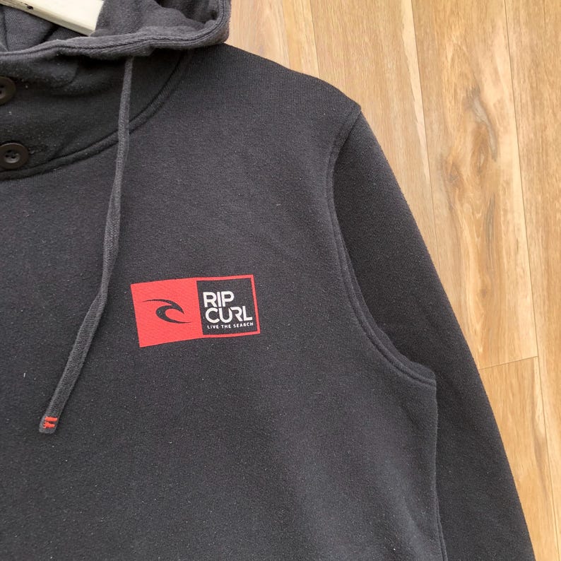 May include: A black hooded sweatshirt with a drawstring and a buttoned neckline. The sweatshirt features a red square logo with the words "RIP CURL" and the tagline "LIVE THE SEARCH" in white.