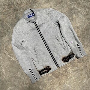 May include: A gray zip-up jacket with a black zipper and two front pockets. The jacket has a stand-up collar and black leather accents with buckles. The sleeves have zippers at the cuffs. The jacket has the word "MAN" on the collar.