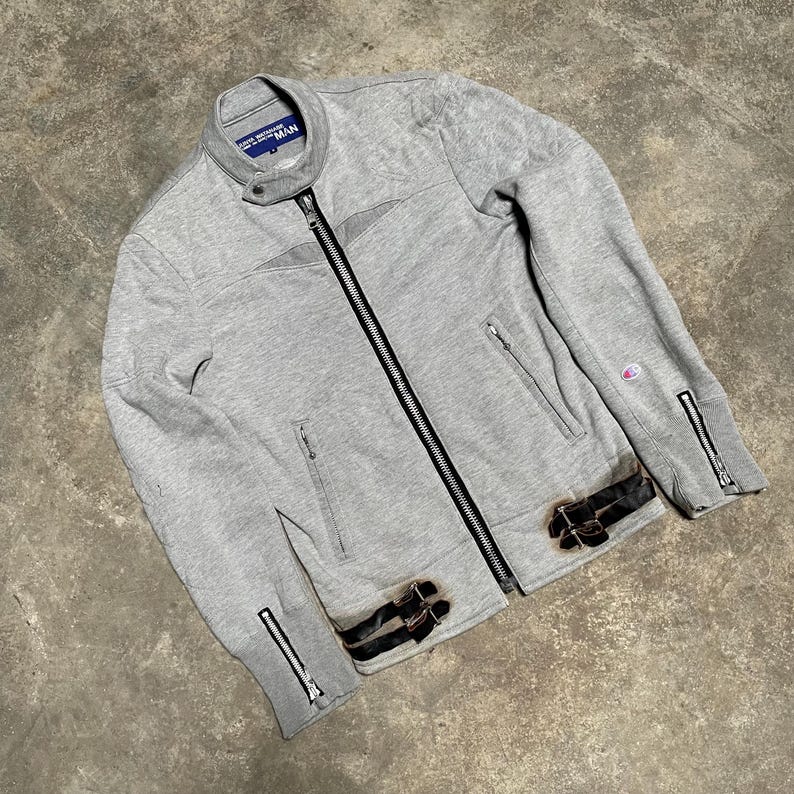 May include: A gray zip-up jacket with a black zipper and two front pockets. The jacket has a stand-up collar and black leather accents with buckles. The sleeves have zippers at the cuffs. The jacket has the word "MAN" on the collar.