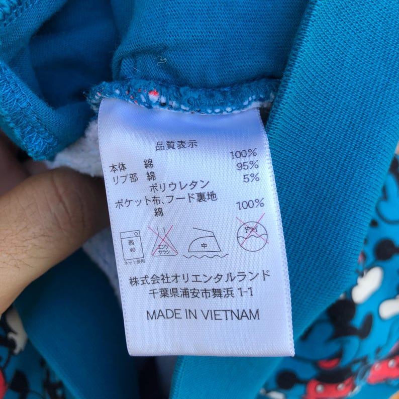 May include: Close-up of a clothing tag with Japanese text and washing instructions. The tag is white with black text and is attached to a blue garment. The tag reads "MADE IN VIETNAM".