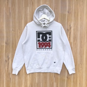 May include: White hooded sweatshirt with a black and red DC Shoes logo featuring the year 1994. The logo is inside a square with a black mesh pattern.