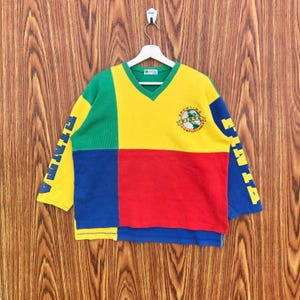 Puede incluir: Una camiseta de manga larga con bloques de color y cuello en V. La camiseta presenta un diseño de patchwork con paneles verdes, amarillos, rojos y azules. Las mangas tienen la palabra "FINTA" impresa en amarillo. Un logotipo con el texto "SPORTING COMFORTABLE" está en el pecho.