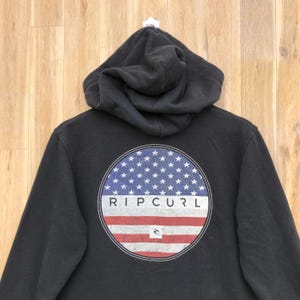 May include: Black hooded sweatshirt with a circular graphic on the back. The graphic features a blue field of white stars above red and white stripes, with the brand name "RIP CURL" in white.