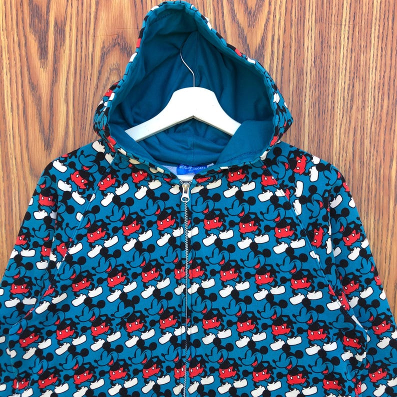 May include: A teal zip-up hoodie featuring a repeating pattern of Mickey Mouse. The design includes red, black, and white Mickey Mouse figures against a teal background. The hoodie has a teal-colored hood and a white hanger.
