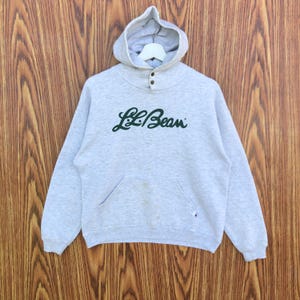 May include: A light gray hooded sweatshirt with the text "L.L.Bean" in green script across the chest. The sweatshirt has a kangaroo pocket, a buttoned neckline, and a hood. The background is a wooden surface.