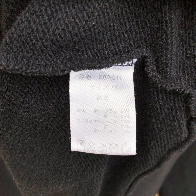 May include: Close-up of a dark gray fabric with a white tag. The tag has Japanese text and details the fabric composition: 63% polyester, 35% cotton, 63% polyester in the ribbing, 33% cotton, and 4% polyurethane.