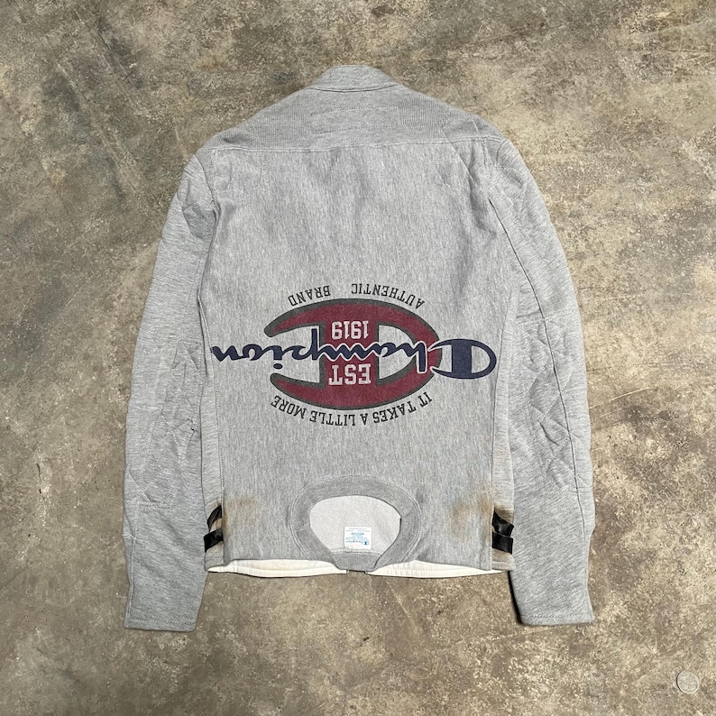 May include: Gray Champion sweatshirt with a quilted back. The Champion logo is in red, navy, and white, with the text "EST 1919" and "IT TAKES A LITTLE MORE". The sweatshirt has a crew neck and long sleeves.