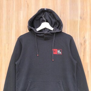 May include: Black hooded sweatshirt with a kangaroo pocket and drawstring hood. Features a small red and white logo on the chest that reads "RIP CURL". The sweatshirt has a button closure at the neck.