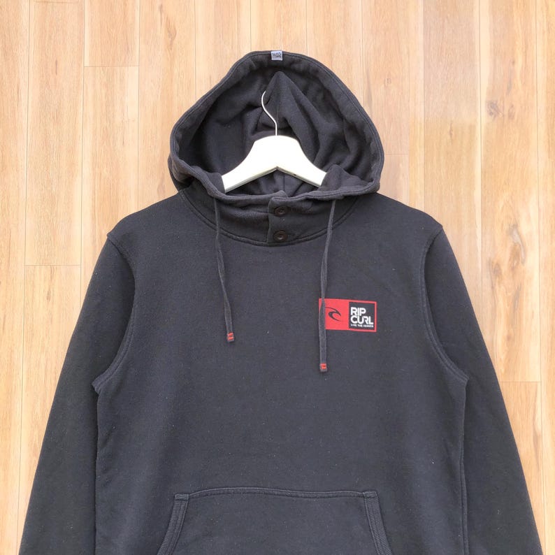 May include: Black hooded sweatshirt with a kangaroo pocket and drawstring hood. Features a small red and white logo on the chest that reads "RIP CURL". The sweatshirt has a button closure at the neck.