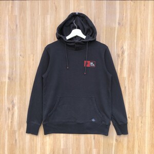 May include: Black hooded sweatshirt with a front pocket and a small logo on the chest. The sweatshirt has a drawstring hood and a button closure at the neck. The Rip Curl logo is in red and white.
