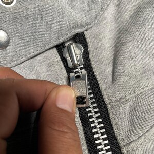 May include: Close-up of a gray fabric with a silver zipper. The zipper is partially closed, revealing the teeth and the YKK brand marking. The fabric appears to be a soft, knit material.