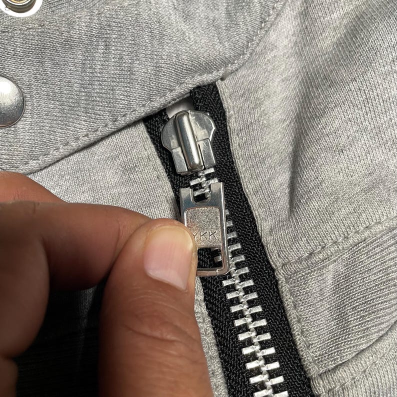 May include: Close-up of a gray fabric with a silver zipper. The zipper is partially closed, revealing the teeth and the YKK brand marking. The fabric appears to be a soft, knit material.