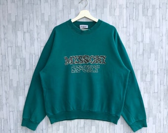 Puede incluir: Sudadera de color verde azulado con el texto "MISSONI SPORT" en una fuente decorativa y texturizada en el pecho. La sudadera está colgada en una percha blanca contra una pared de ladrillos gris claro.