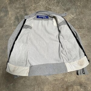 May include: A gray zip-up jacket with a light gray interior and black zippers. The jacket has a small blue tag with white text. The jacket has two front pockets and white accents.