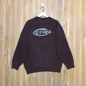 May include: A dark brown sweatshirt with the word "GESCHWA" printed in a light blue oval shape on the back. The sweatshirt is hanging on a white hanger against a light brown wooden background.