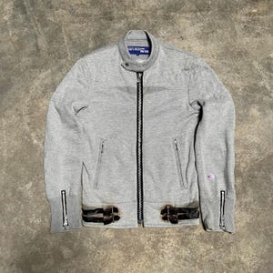 May include: A light gray zip-up jacket with a stand-up collar and black accents. The jacket features two front pockets, a full-length zipper, and black leather-like straps with buckles at the waist. The label reads "JUNYA WATANABE MAN".