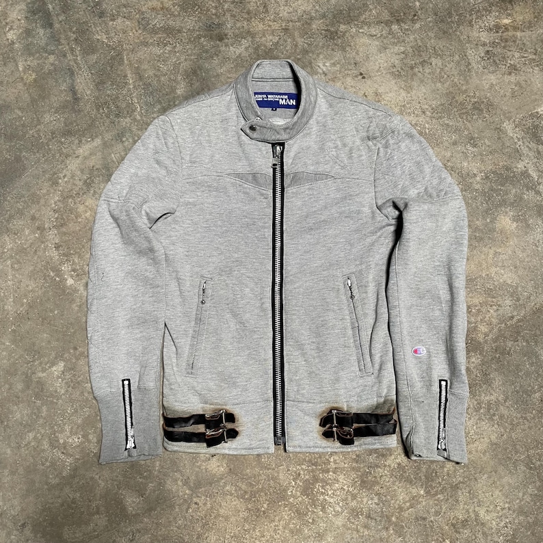 May include: A light gray zip-up jacket with a stand-up collar and black accents. The jacket features two front pockets, a full-length zipper, and black leather-like straps with buckles at the waist. The label reads "JUNYA WATANABE MAN".