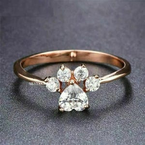 Può includere: Un anello in oro rosa con una pietra centrale trasparente a forma di cuore e pietre trasparenti più piccole disposte a forma di impronta di zampa.
