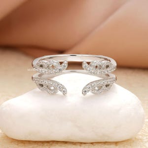 May include: A silver-toned ring with a unique design. The ring features two curved bands, each adorned with small, sparkling clear stones. The bands curve around the finger, creating an open, modern look. The ring is set on a white stone.
