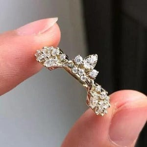 Könnte beinhalten: Ein goldfarbener Ring mit floralem Design, besetzt mit mehreren klaren Edelsteinen. Der Ring wird zwischen zwei Fingern gehalten, wodurch seine komplizierten Details und sein Glanz hervorgehoben werden. Das Design umfasst Cluster aus kleinen Steinen und größere, blattförmige Edelsteine.