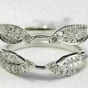 Pode incluir: Dois realçadores de anel prateados, cada um com um design curvo com pequenas pedras preciosas transparentes. Os realçadores são projetados para acomodar uma pedra central. Os anéis estão em um fundo branco.