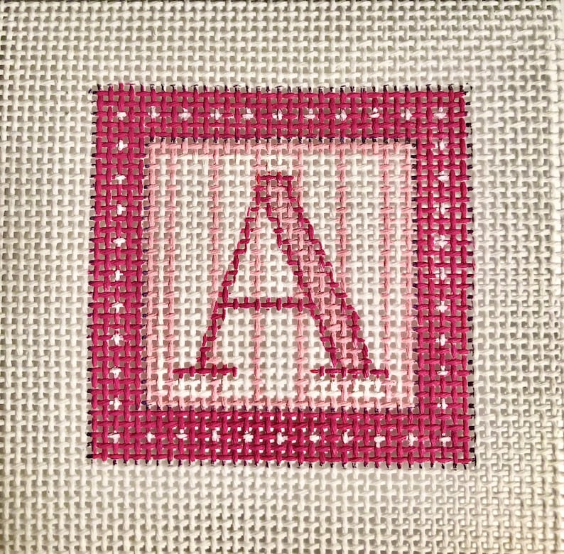 May include: A hand-stitched needlepoint canvas featuring the letter "A" in a pink and white color scheme. The letter is centered within a square frame, with a dotted border. The background is a neutral, off-white color.