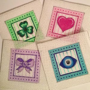 May include: Four square needlepoint canvases with different designs. One has a green shamrock, another a pink heart, a third a purple bow, and the last a blue eye. Each design is framed with a contrasting color.