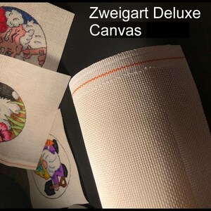 May include: A close-up of a roll of Zweigart Deluxe Canvas with an orange thread border. Several small, round canvases with colorful embroidered designs are visible, including a bunny, a gnome, and a cat.