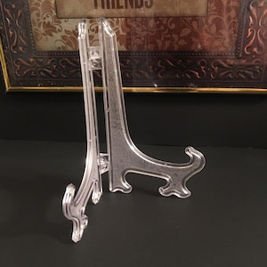 May include: Clear plastic display easel with a curved base and a vertical support. The easel is designed to hold a framed picture or other decorative item. The easel is made of clear plastic.