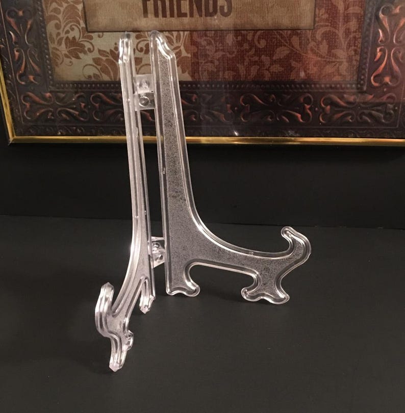 May include: Clear plastic display easel with a curved base and a vertical support. The easel is designed to hold a framed picture or other decorative item. The easel is made of clear plastic.