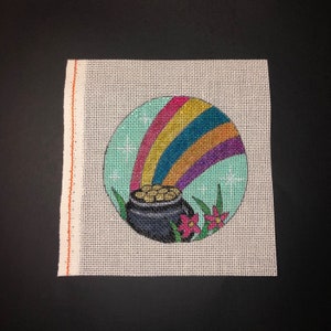 May include: A needlepoint canvas with a round design of a pot of gold at the end of a rainbow. The rainbow is multi-colored with red, orange, yellow, green, blue, indigo, and violet stripes. The pot of gold is black with gold coins inside. There are green leaves and pink flowers around the pot of gold.