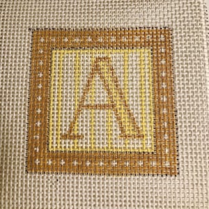 May include: Needlepoint canvas featuring the letter "A" in a square frame. The letter and frame are in shades of yellow and brown, with a dark brown border. The background is a neutral, off-white woven fabric.