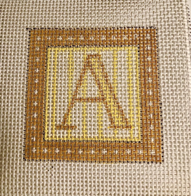 May include: Needlepoint canvas featuring the letter "A" in a square frame. The letter and frame are in shades of yellow and brown, with a dark brown border. The background is a neutral, off-white woven fabric.