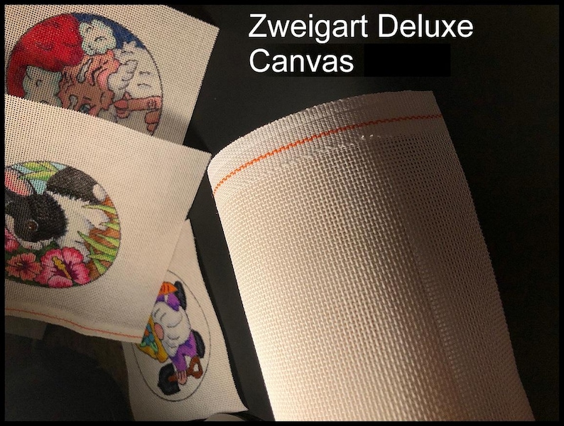 May include: A close-up of a roll of Zweigart Deluxe Canvas with an orange thread border. Several small, round canvases with colorful embroidered designs are visible, including a bunny, a gnome, and a cat.