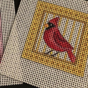 May include: Needlepoint canvas featuring a red cardinal bird within a yellow square border. The bird is detailed with black accents and sits on a thin black line. The background is a white, open-weave fabric.