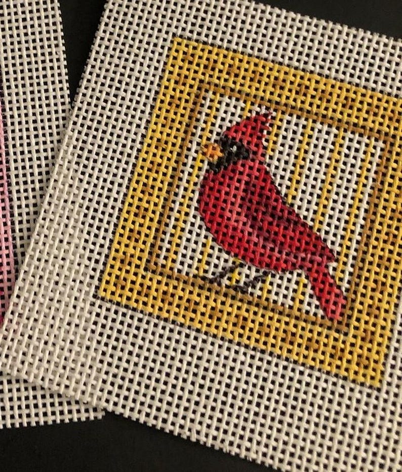 May include: Needlepoint canvas featuring a red cardinal bird within a yellow square border. The bird is detailed with black accents and sits on a thin black line. The background is a white, open-weave fabric.